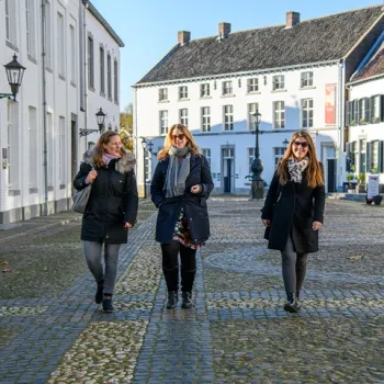 Drie vrouwen in winterkleding wandelen lachend over een zonnig plein met kinderk